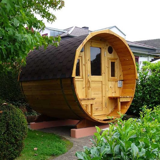 A barrel-shaped outdoor sauna with a glass door and outdoor seating area, placed on a grassy area beside some bushes.