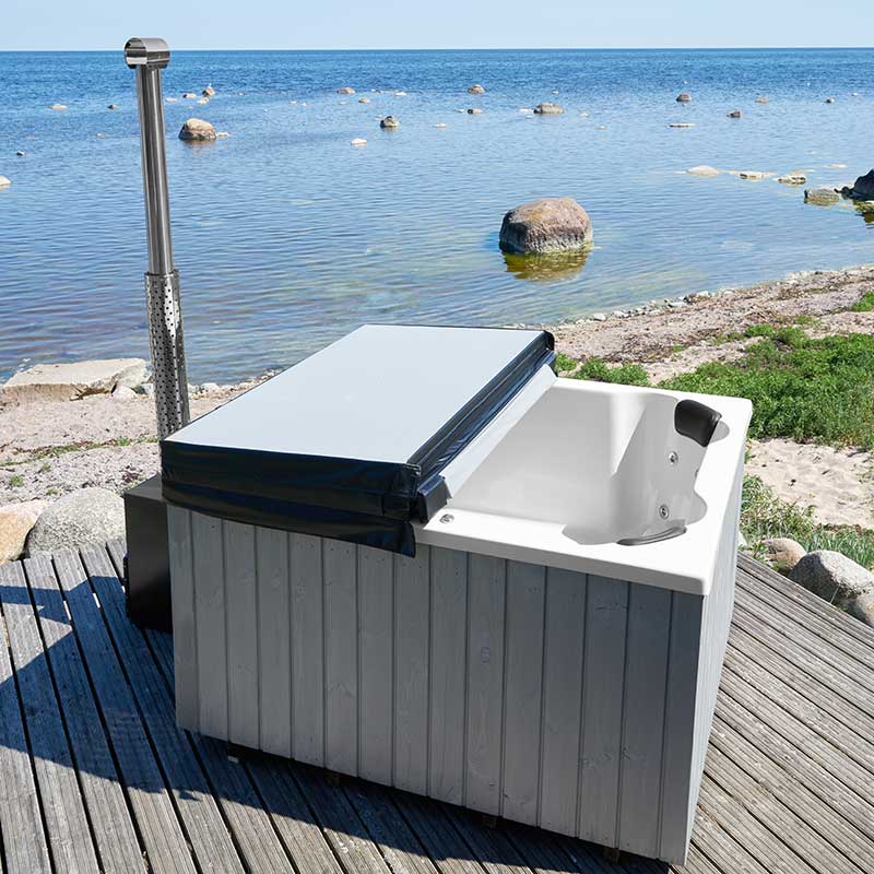 A square, fiberglass hot tub with wood-fired heating, positioned on a wooden deck with an external wood stove, by a body of water.