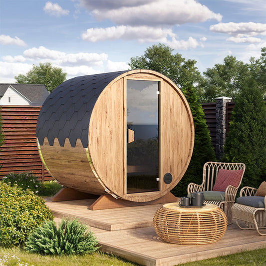 A compact outdoor barrel sauna for 2-3 persons, placed on a wooden platform with outdoor furniture around. The sauna has a brown wooden exterior, a tempered glass door, and a bitumen shingle roof.