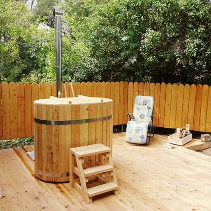 A compact oval-shaped wooden hot tub with an exterior heater on a wooden deck, surrounded by lush greenery. A pair of wooden steps leads to the hot tub, and there is a lawn chair beside it.