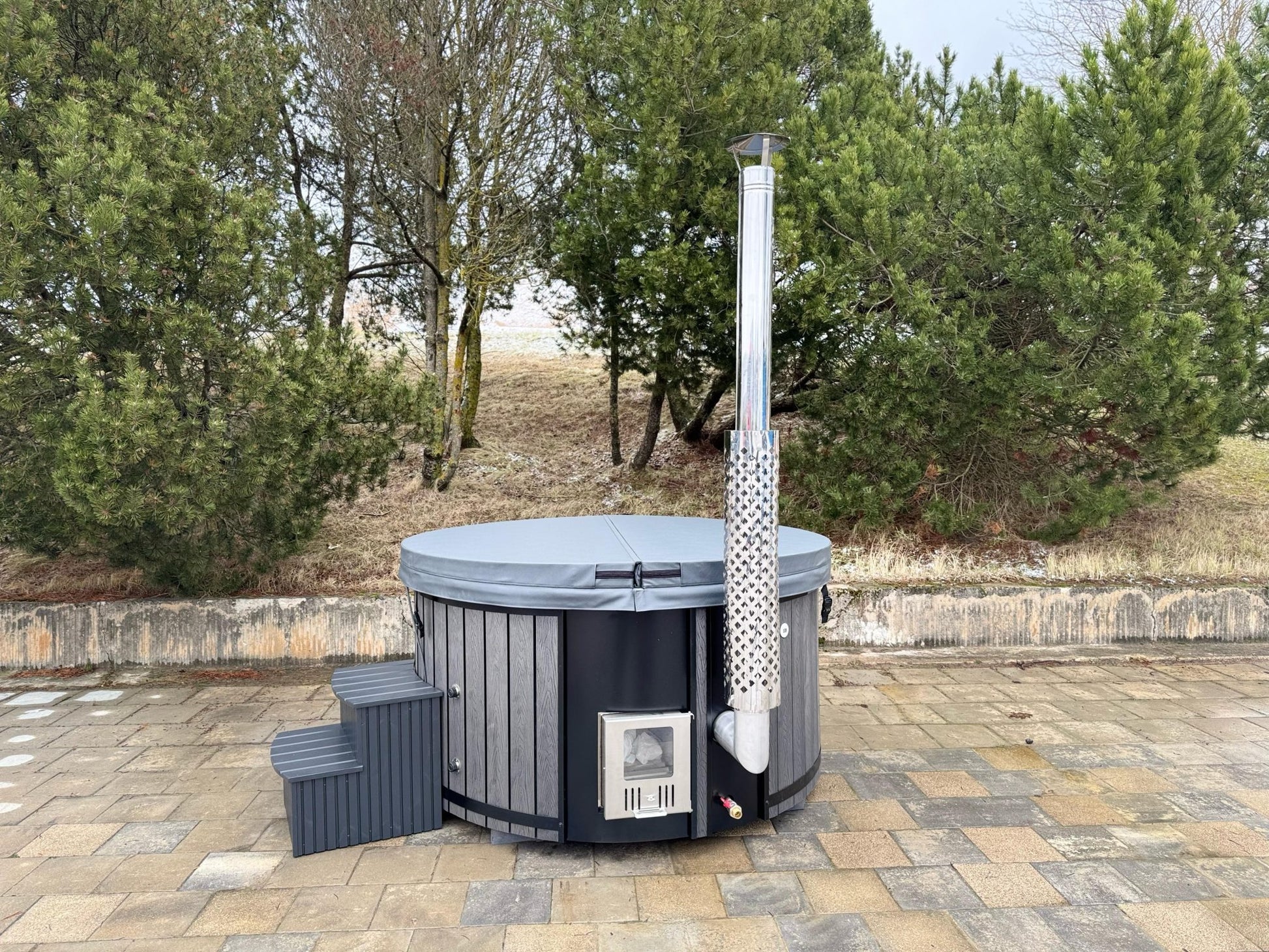 Hot tub with a chimney-like structure outdoors on a paved area with trees in the background