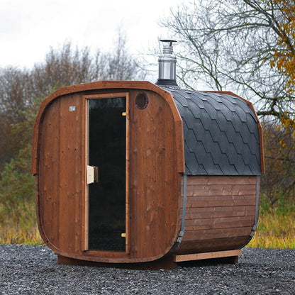 A brown cube-shaped barrel sauna with a glass door and a shingle roof, set outdoors.